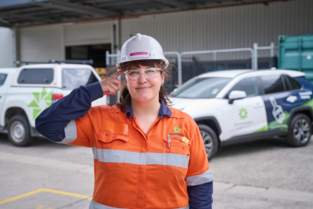 A person in an orange safety uniform and hard hat stands outdoors, pointing to their safety glasses, with two vehicles parked behind them.