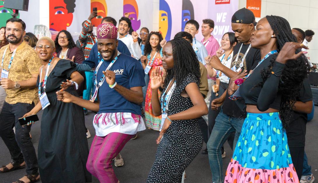 A diverse group of people joyfully dancing together indoors, wearing colorful clothing and lanyards