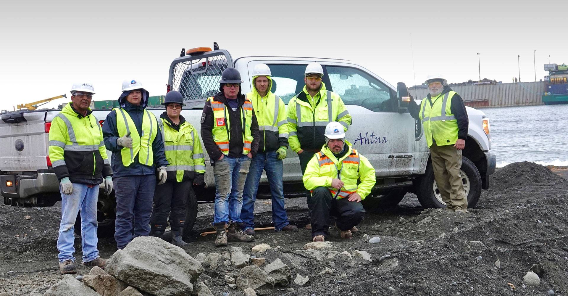 A group of construction workers in high-visibility jackets and helmets stand next to a pickup truck near a waterfront, with rocky terrain in the foreground.