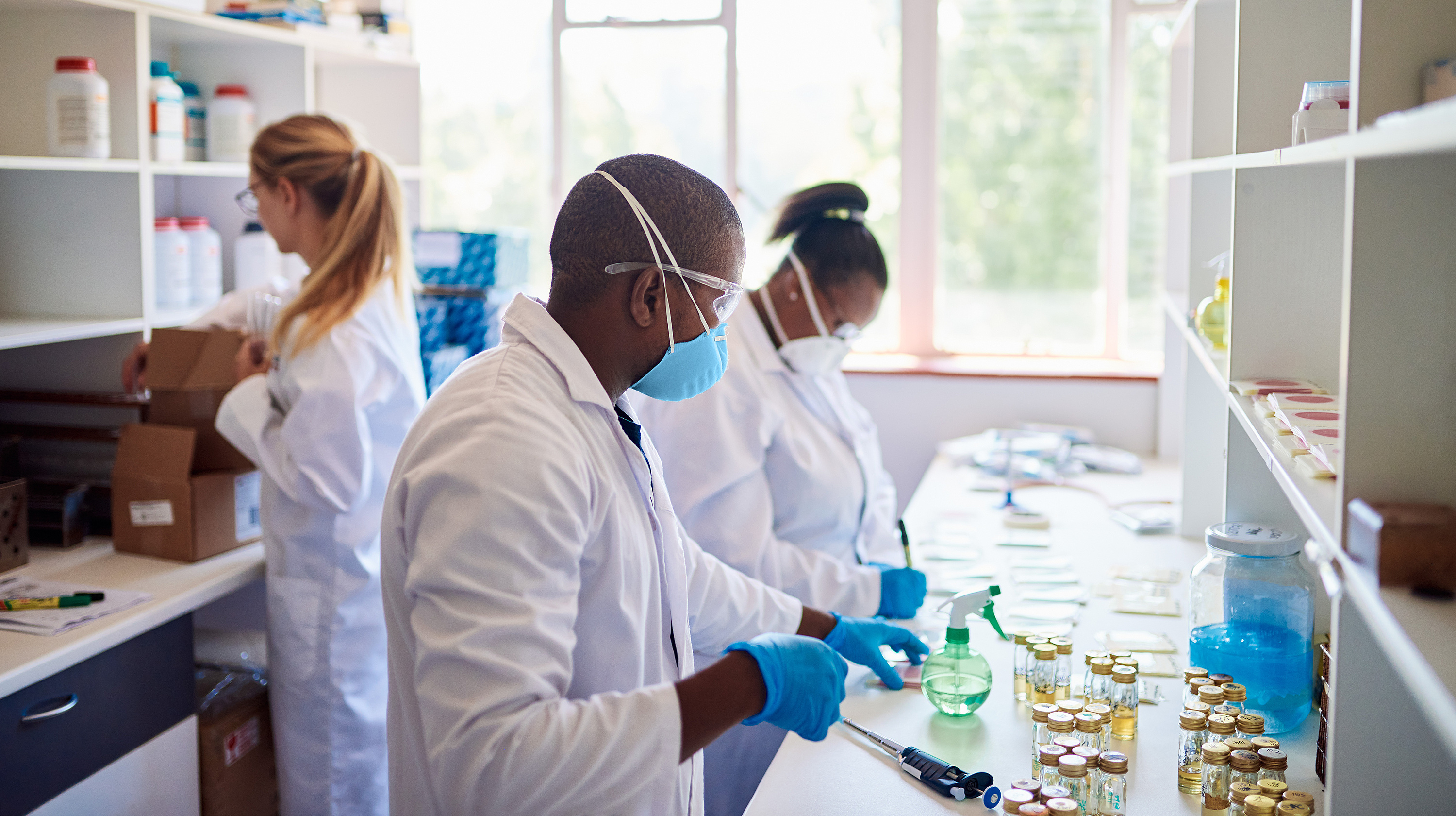 Three scientists in lab coats and masks work in a bright laboratory